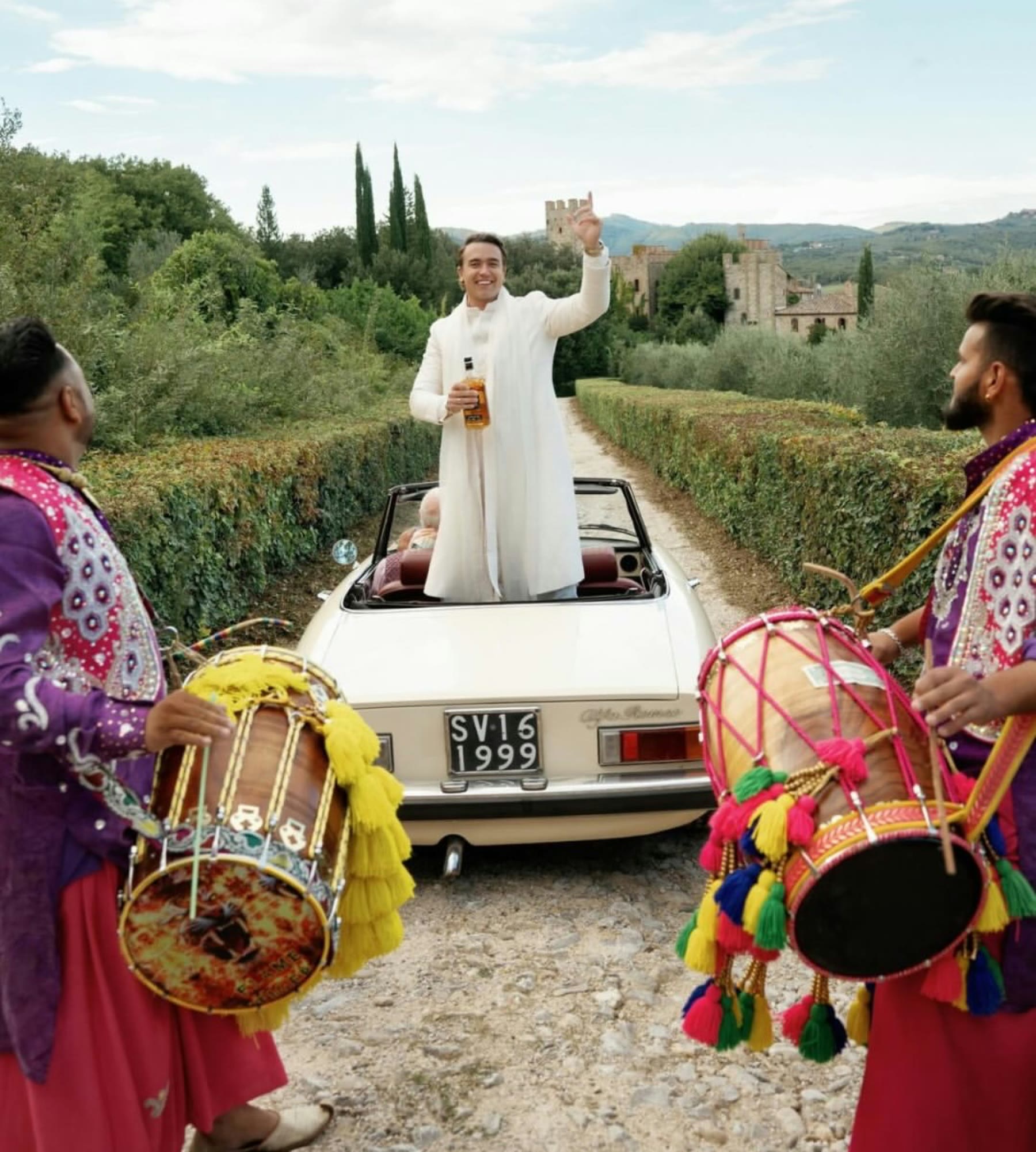 Nick in piedi su un'auto d'epoca scoperta, fra due suonatori di dhol